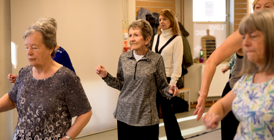 A class of women dancing in a group, the woman in the middle has short hair with a grey zip-up top and black leggings
