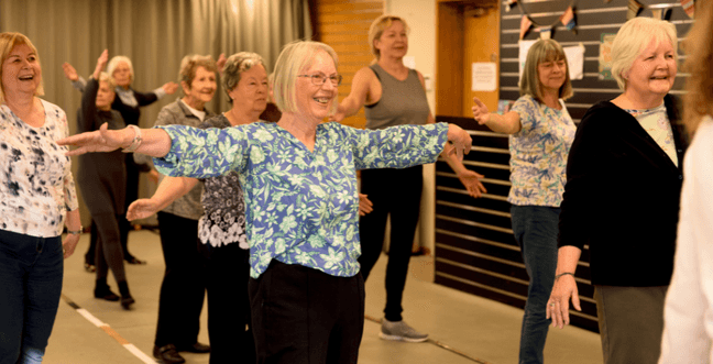 A woman with blonde short hair and glasses with her arms outstretched wears a blue shirt and black trousers in a dance class