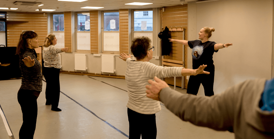 A group of people in a dance class hold their arms raised by their sides