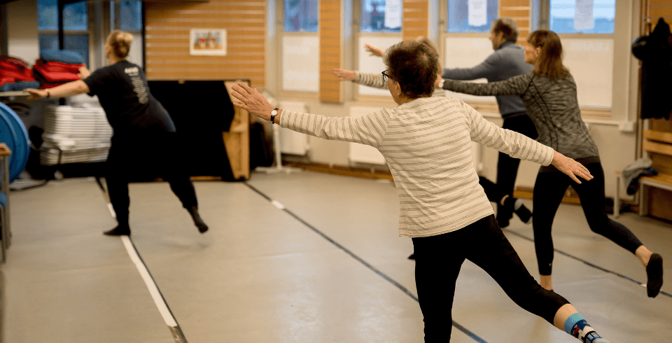 A group of people in a dance class stand on one leg with arms outstretched