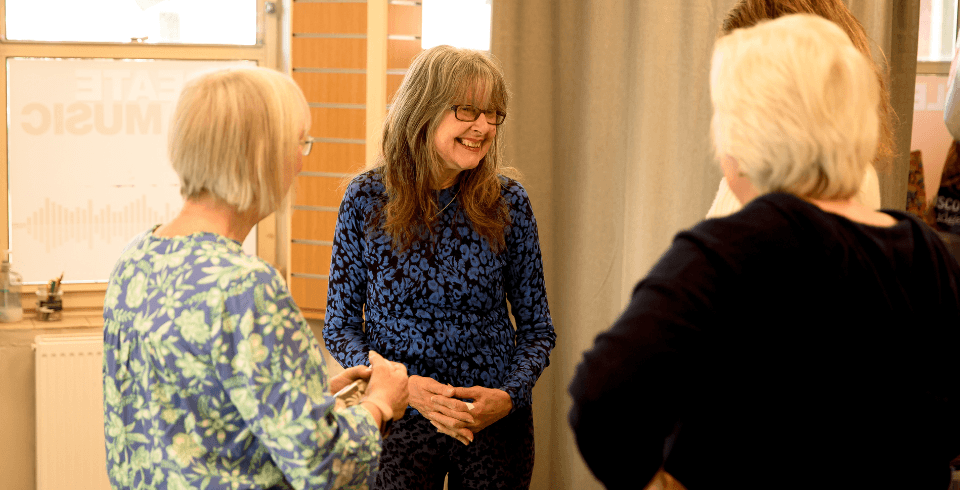 Three women talking in a group, the woman in the middle smiles with grey long hair, a fringe, black square glasses and a blue top