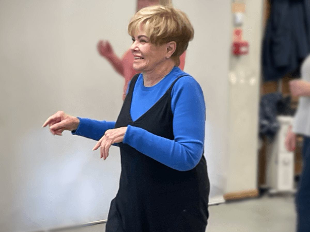 A white woman in a blue top and black jumpsuit with blonde short hair, smiles as she dances on the spot
