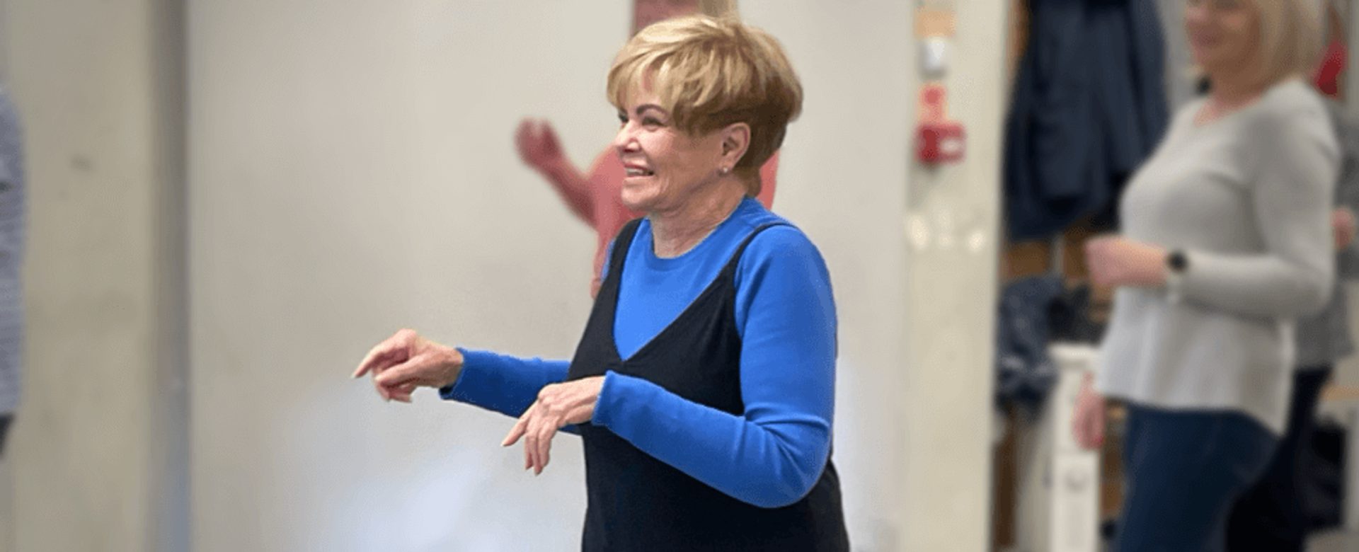 A white woman in a blue top and black jumpsuit with blonde short hair, smiles as she dances on the spot