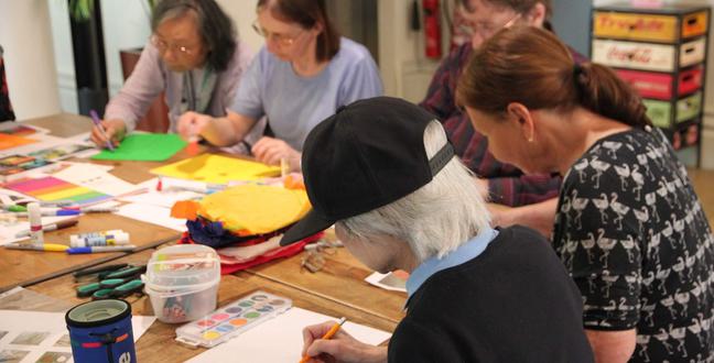 A group of women sat at a wooden table with art supplies laid out on the table
