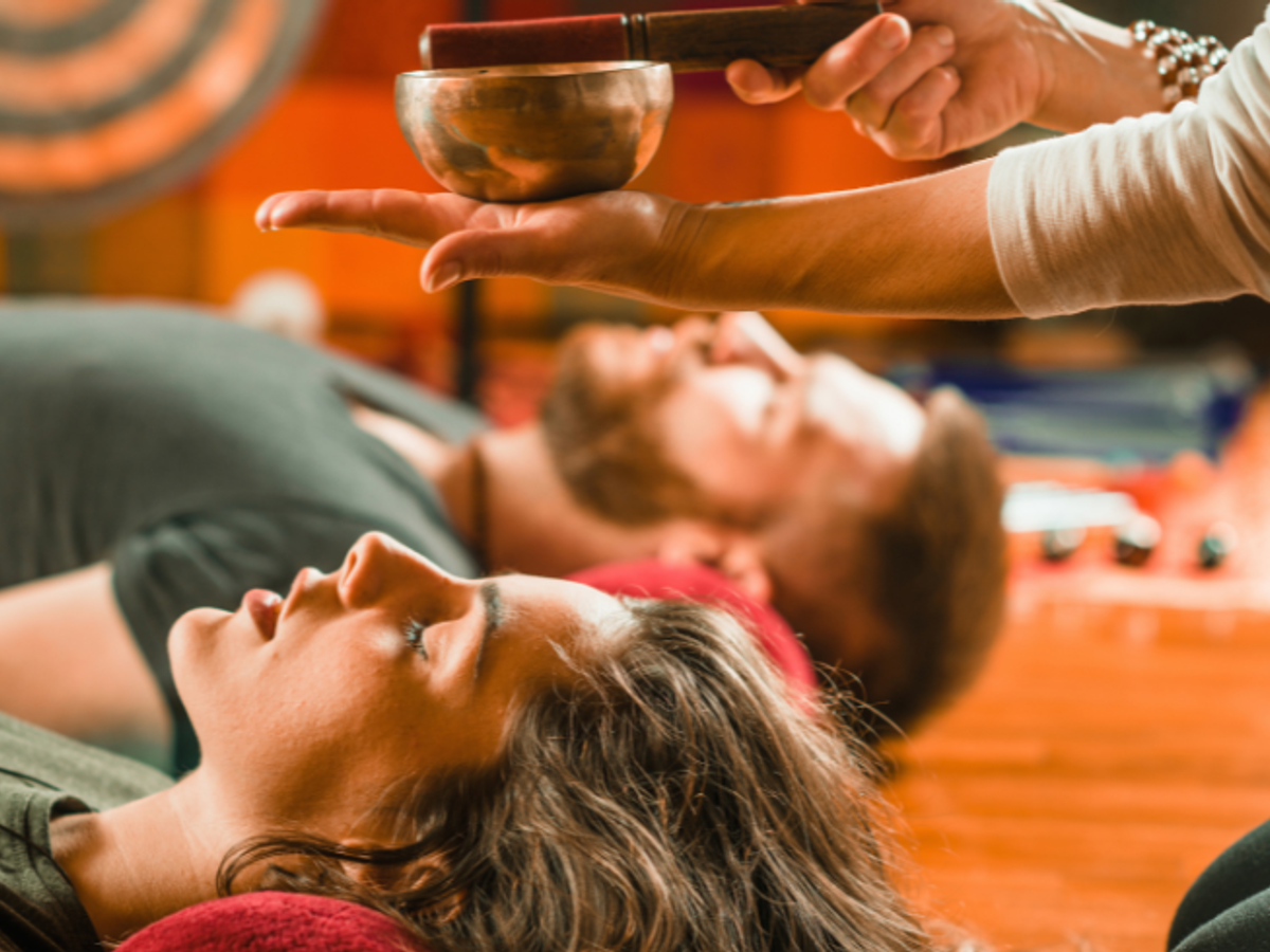 A man and a women lead on the floor. A metal gong can be seen in the background. Above the man and women's heads someone holds a metal bowl and uses a wooden stick to play it in the palm of their hand.