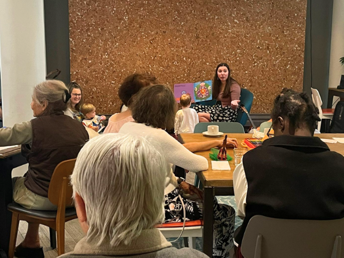 A group of adults sit and watch a woman with long brown hair read a picture book to a group of toddlers