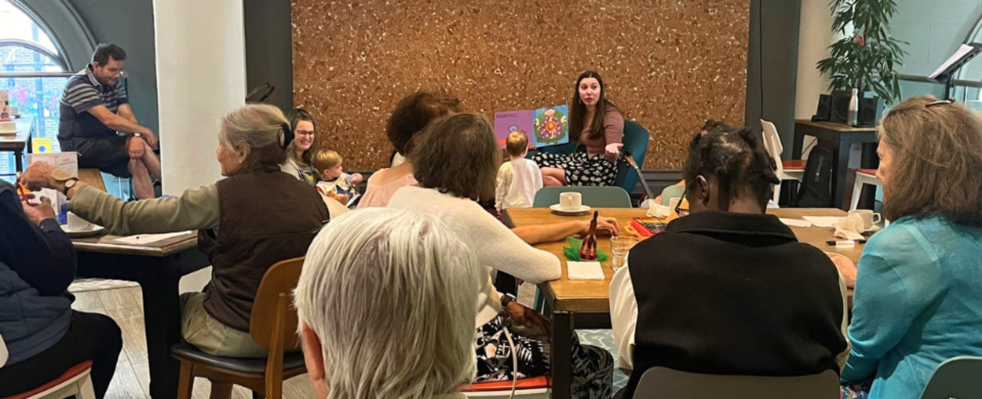 A group of adults sit and watch a woman with long brown hair read a picture book to a group of toddlers