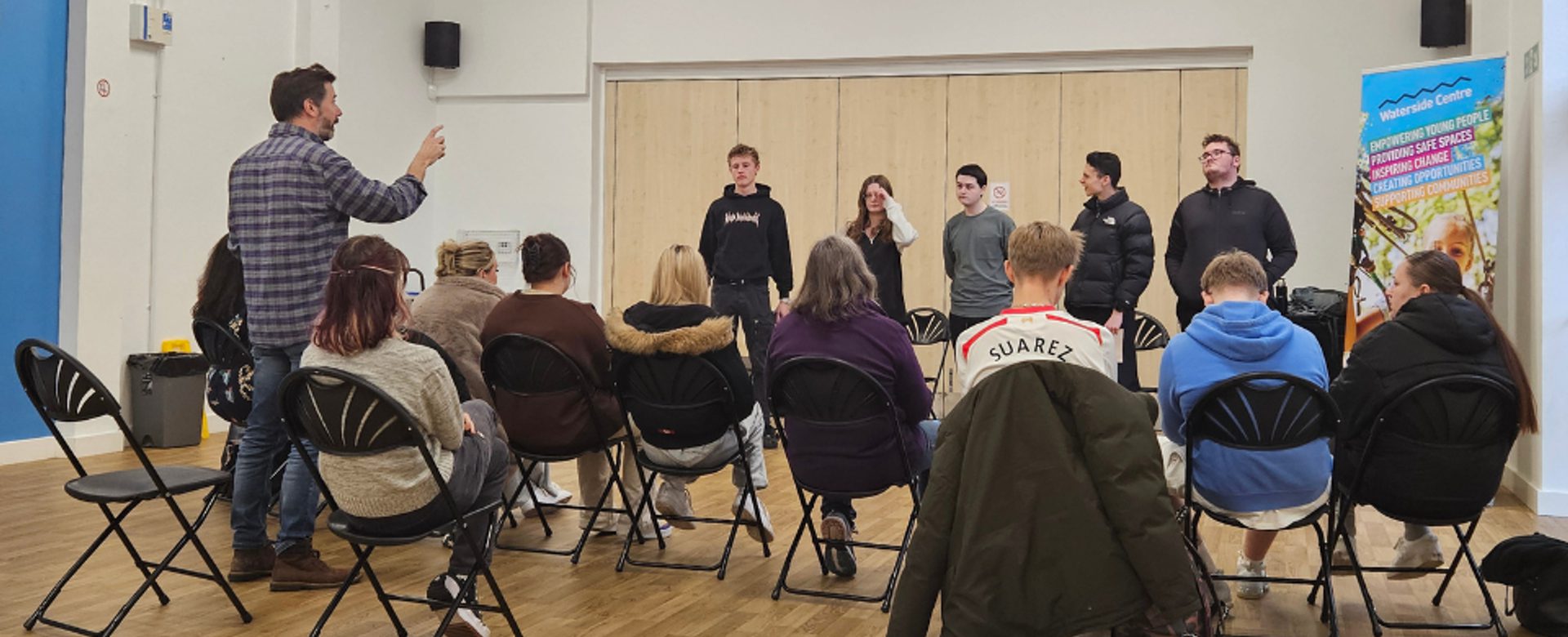 A group of students stand in a hall in front of people sat on black fold up chairs while a man in the crowd stands up pointing a finger