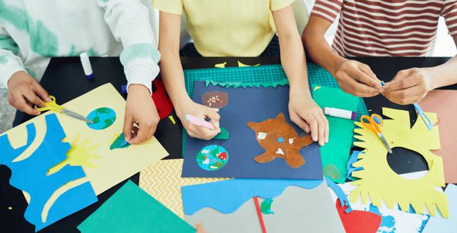Three children's hands crafting with multi-coloured paper at a desk