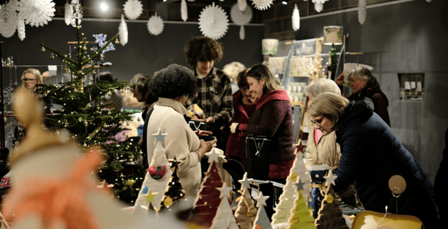 Paper snowflakes hang from the ceiling of the gallery space, in the centre of the room is a Christmas tree and people can be seen chatting and looking at the various stalls
