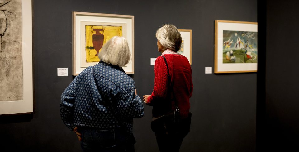 Two people looking at paintings on a dark wall, one in a blue jacket and the other in a red cardigan