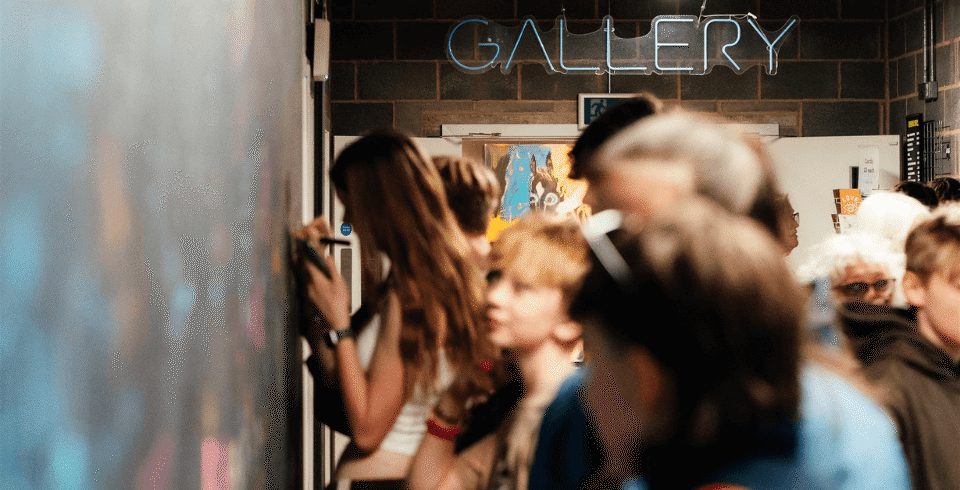 A large group of people drawing on a black wall with the neon sign for a gallery behind them