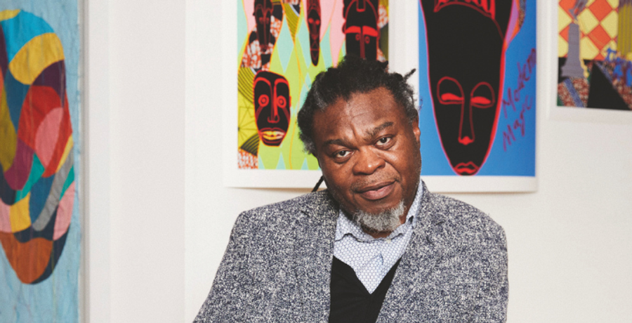 A black man in a grey suit with a blue shirt underneath sits in front of large colourful artworks on a white wall