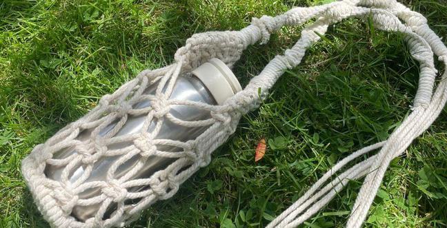 A silver flask lies on the grass while sat in a cream macramé holder with a long handle
