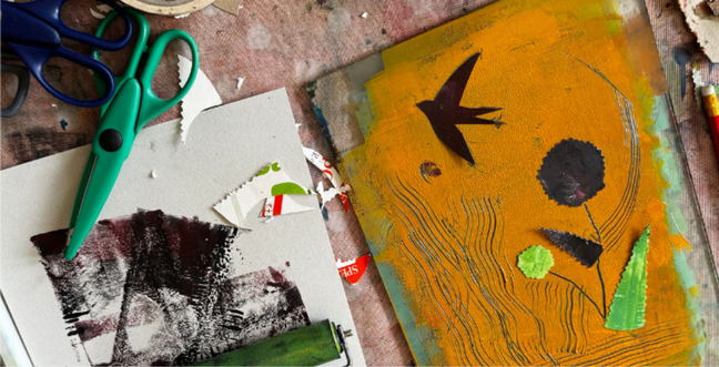 A wooden table with printing materials and green scissors. The print in the centre is a bird and flower shadow on orange card