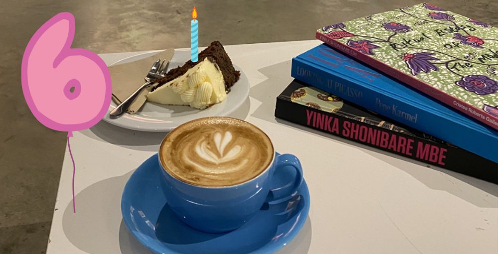 A coffee in a blue mug with latte art sits on a white table next to a cake with an animated candle and a balloon in 6