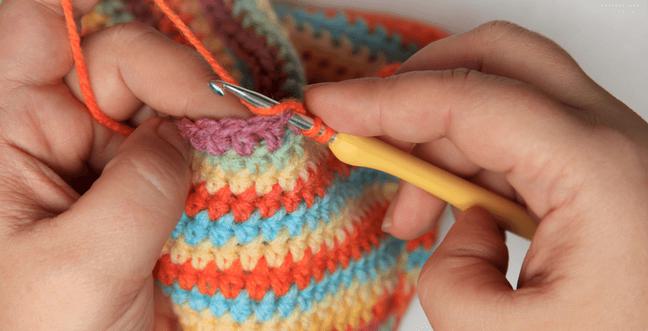 A person uses a silver crochet hook with a yellow handle to create a rainbow piece of crochet