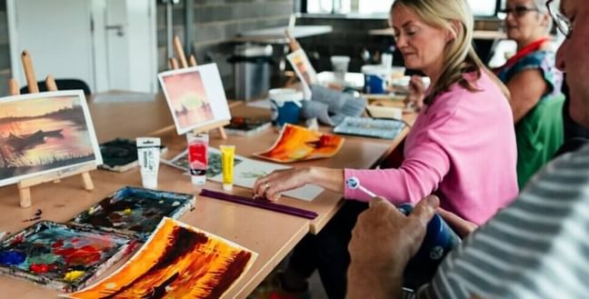 A group of people sit at a long wooden table in front of mini easels with sunset pictures on them while they paint a sunset on white paper