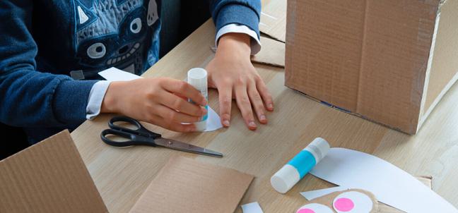 A young persons hands wearing a blue jumper with a monster on the front uses a glue stick on white paper sat at a wooden table with cardboard, glue sticks, paper and scissors