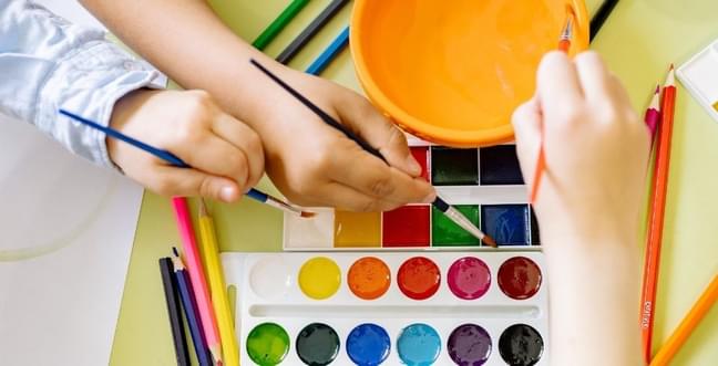 Children's hands over a paint palette with paint brushes and an orange bowl