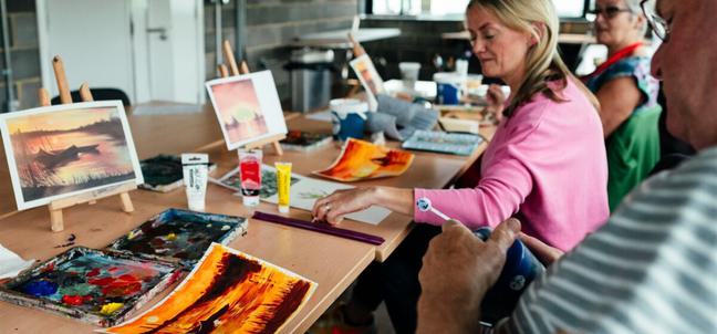 A group of people sit at a long wooden table in front of mini easels with sunset pictures on them while they paint a sunset on white paper