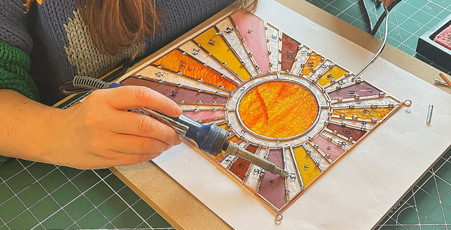 A person using a soldering iron to solder together glass pieces lined with copper wire. The glass create the image of a gleaming sun