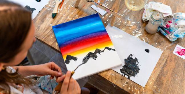 A ariel view of a woman painting on a canvas with a rainbow gradient on a wooden table with a glass of wine
