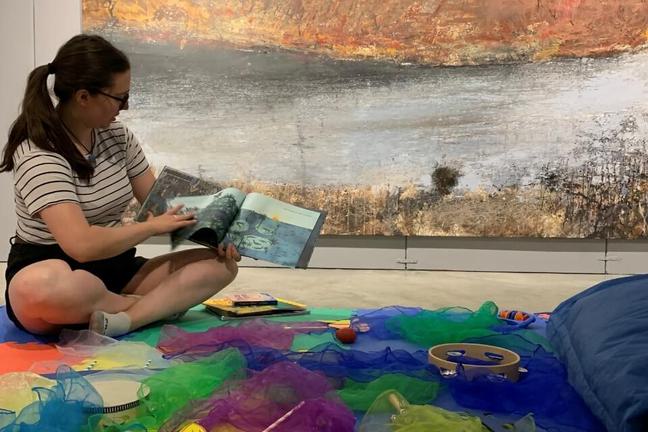 A woman with brown hair and glasses sits on the gallery floor on a playmat, reading a storybook