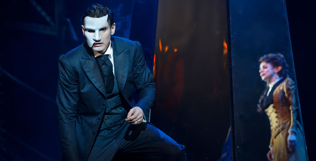 A man wearing a white broken mask and a woman stands to the side behind him.