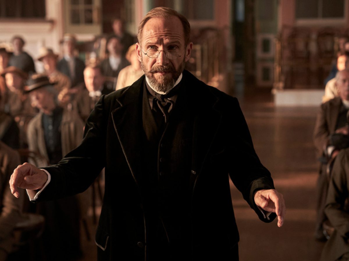A man wearing an old-fashioned suit stood in the middle of a hall with lost of other people.