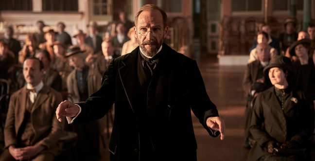A man wearing an old-fashioned suit stood in the middle of a hall with lost of other people.