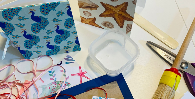 A collection of items including two patterned notebooks, one with peacocks and one with shells on a white desk with ribbon and scissors