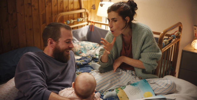 A woman speaking through sign language to a man and a baby as they all sit on a bed.