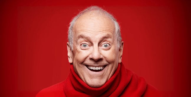 An older man smiling widely and wearing a red jumper against a bright red backdrop.