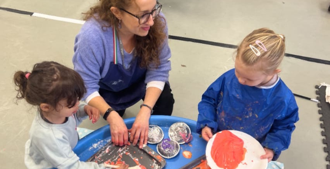 Two little girls sit with a woman as they paint together on a small table