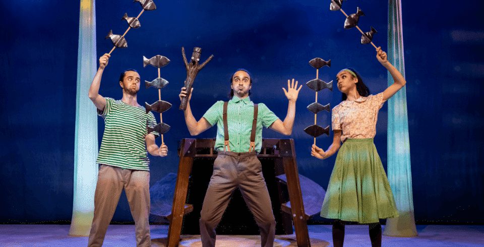 Three performers on a stage hold fish puppets and a wooden Stick Man, all pretending to be underwater.
