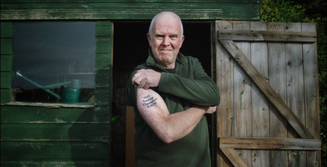 A man holding up his sleeve to show a tattoo as he stands in front of a green shed.