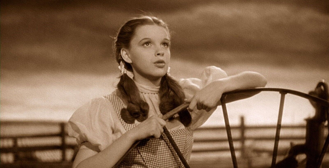 A black and white image of a girl dressed in a gingham dress and looking up at the sky.