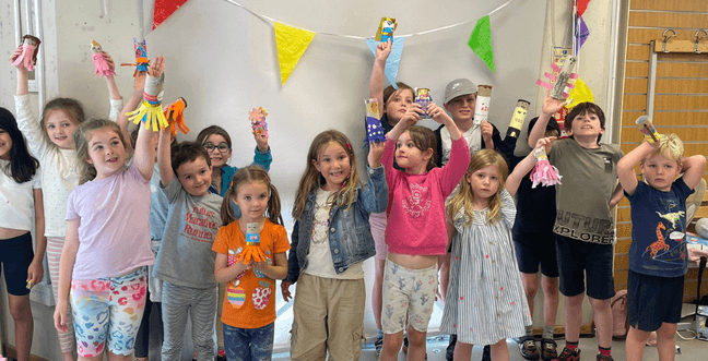 A group of children standing together, proudly holding up colourful craft projects they’ve made, such as decorated paper rolls and other handmade items.