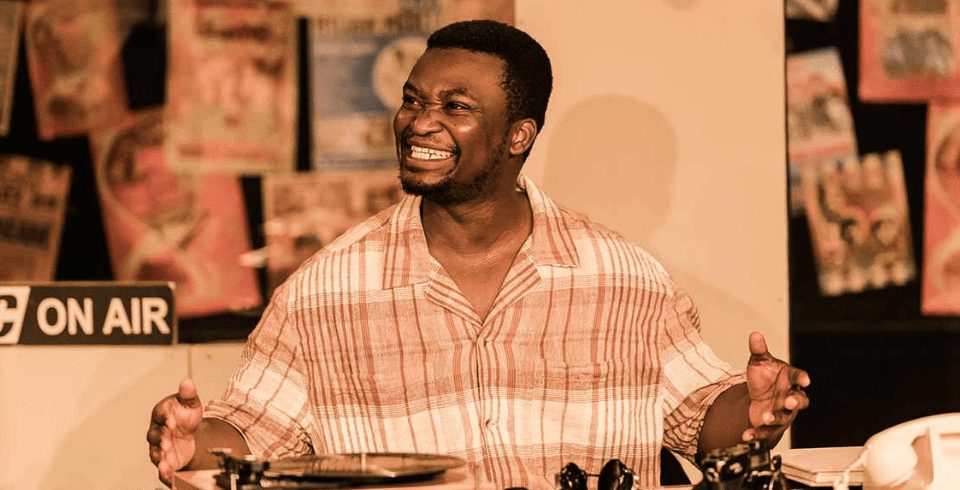 A man in a beige shirt smiling and sat in front of a record player.