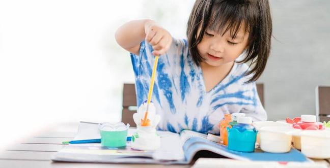 A little girl with dark hair and a fringe sits at a table with a tie dye t-shirt painting