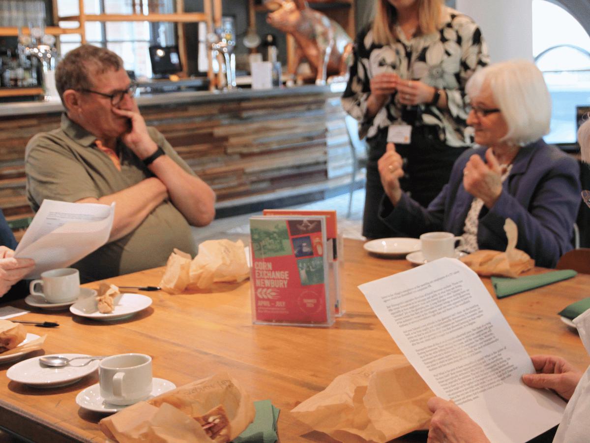 A group of people reading from paper sit around a wooden table in a café with sandwiches in brown bags and hot drinks in white china cups and saucers