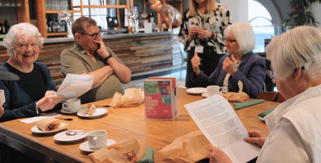 A group of people reading from paper sit around a wooden table in a café with sandwiches in brown bags and hot drinks in white china cups and saucers